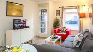 Un salon avec un canapé gris, un fauteuil rouge, un meuble blanc sur lequel est posée une télévision diffusant l’émission « HUSTLE », et une fenêtre donnant sur une voiture garée à l’extérieur.