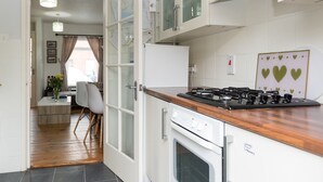 Une cuisine moderne avec des meubles blancs, une cuisinière à gaz et un four. On aperçoit un coin repas avec une table et des chaises à travers une porte ouverte.