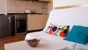 Eine moderne Küche mit Kühlschrank, Backofen und Mikrowelle. Ein weißes Bett mit bunten Kissen tritt in den Vordergrund.