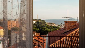 Vista desde una ventana con cortina decorativa, con vistas a un paisaje urbano con tejados de tejas rojas, un edificio de ladrillo y una masa de agua distante.