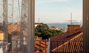 Une vue depuis une fenêtre ornée d’un rideau décoratif, donnant sur un paysage urbain avec des toits recouverts de tuiles rouges, un bâtiment en briques et une étendue d’eau au loin.