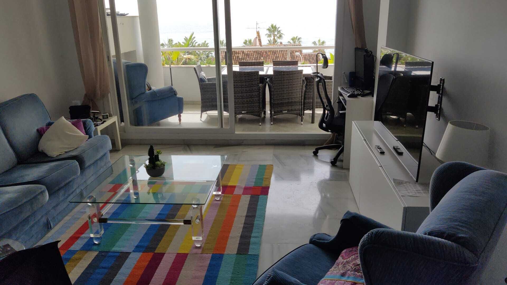A modern living room with a sofa, a coffee table, and a TV. There is a balcony with a view of the sea and palm trees.