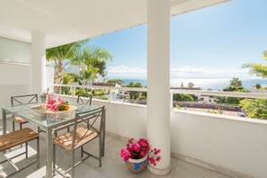 A balcony with a glass table and chairs, a potted plant, and a view of the sea.