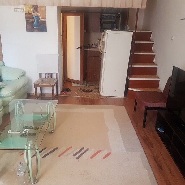 A living room with a glass coffee table, a sofa, a television, and a kitchen area with a refrigerator and microwave.