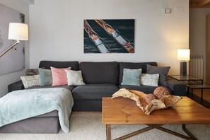 A modern living room with a grey sofa, multiple cushions, a wooden coffee table with decorative items, and a painting of a boat on the wall.