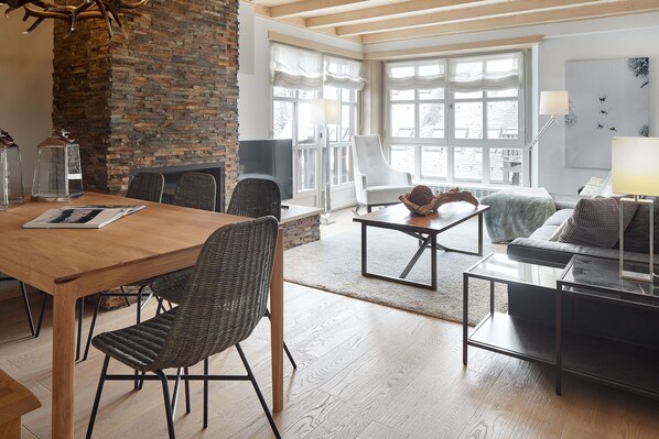 A modern living room with a wooden dining table, wicker chairs, a stone fireplace, and large windows.