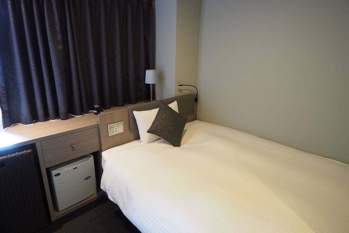 Habitación de hotel con cama, escritorio con lámpara, un mueble pequeño y ventana con cortinas.
