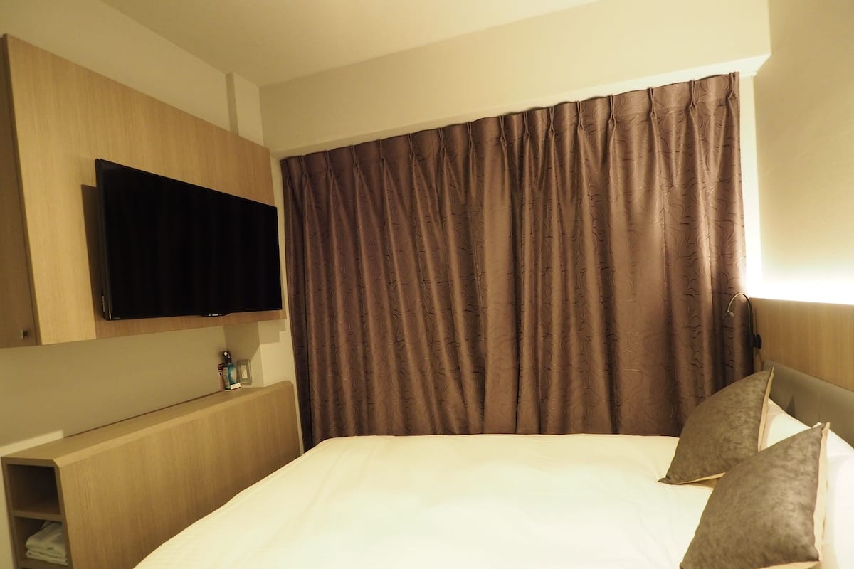 Habitación de hotel con cama, televisor de pantalla plana en la pared, mesita de noche de madera y un ventanal con cortinas.