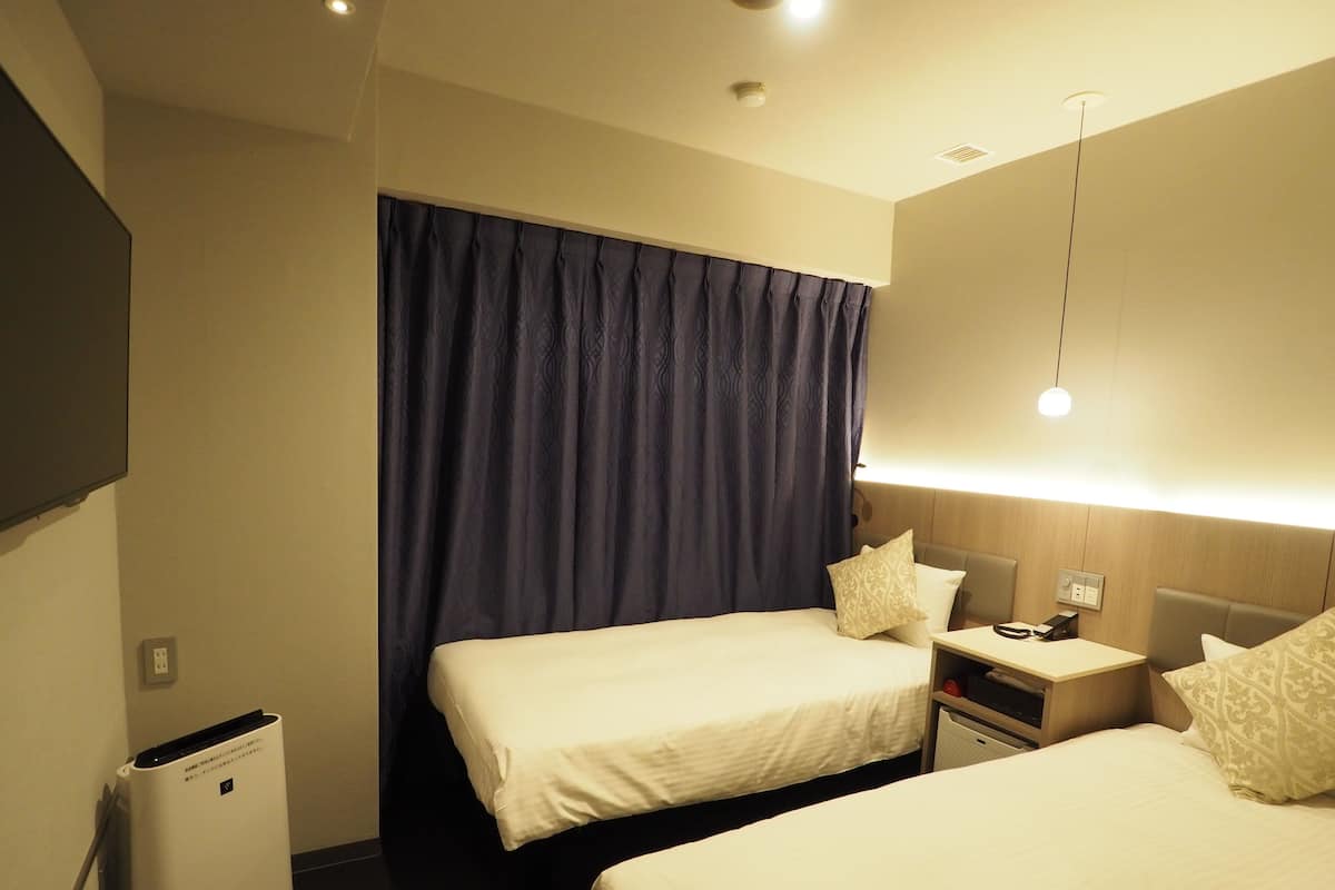 Habitación de hotel con dos camas, una mesita de noche con lámpara, un pequeño calentador y un televisor montado en la pared.