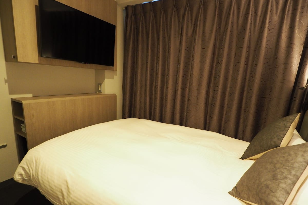 A hotel room with a bed, a television mounted on the wall, and a curtain.