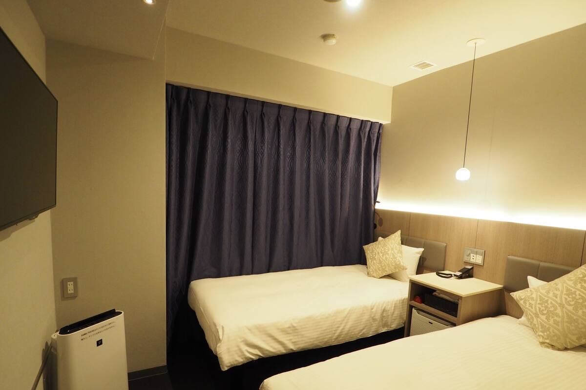 Habitación de hotel con dos camas, una mesita de noche con lámpara, un pequeño calentador y un televisor montado en la pared.