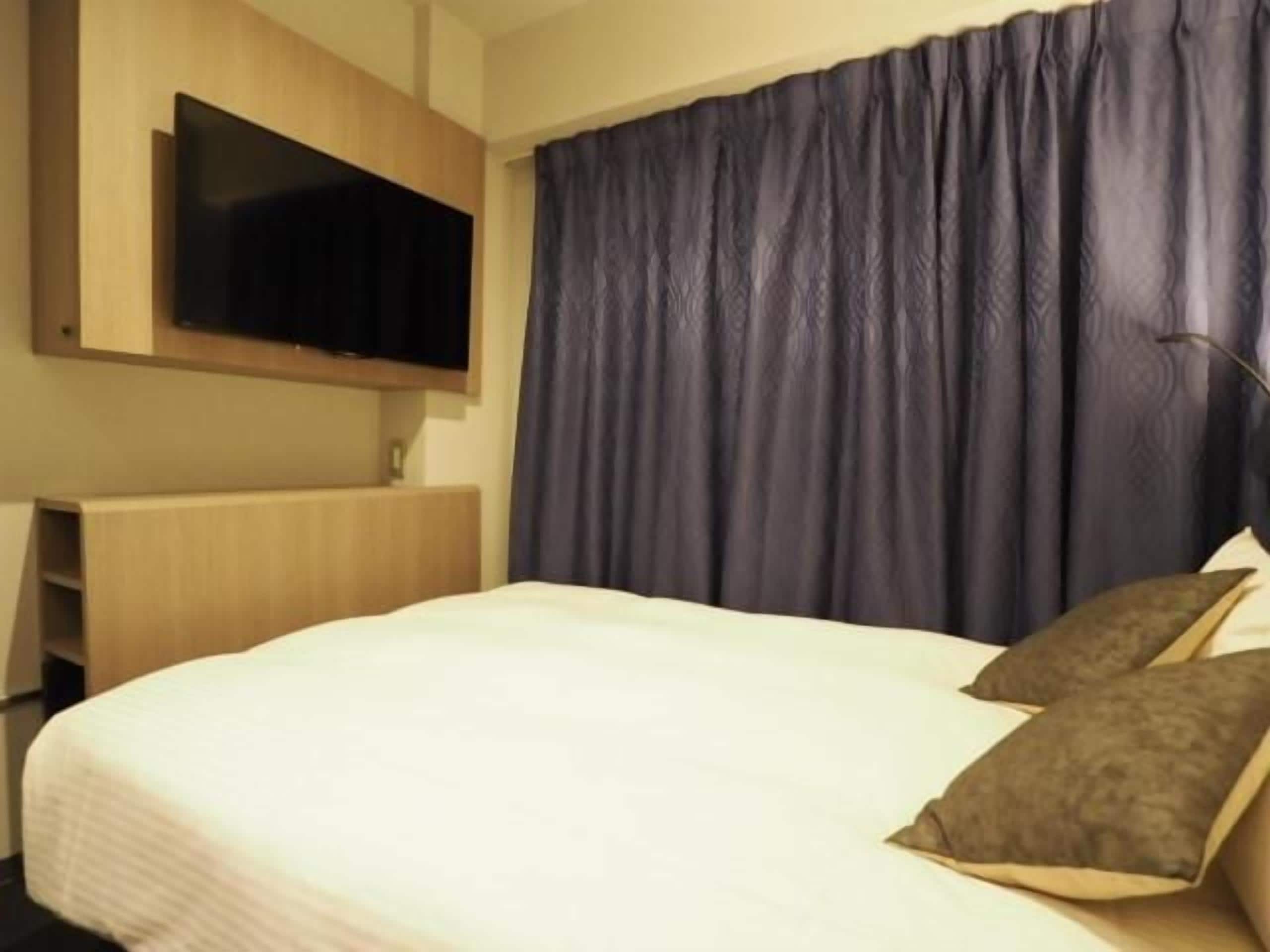 A hotel room with a bed, a flat-screen TV mounted on the wall, and a dark curtain.
