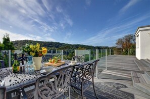 A dining table with chairs, a vase with flowers, bottles, and a fruit plate on a deck with a view of hills. - 6 Bed in Lee (oc-highvi) (Lee)