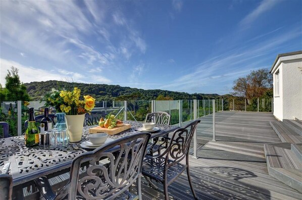 A dining table with chairs, a vase with flowers, bottles, and a fruit plate on a deck with a view of hills.