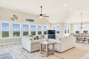 A spacious living room with a large window, a ceiling fan, and a flat-screen TV.