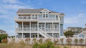 Una casa fronte spiaggia su due piani, con un balcone ad ogni piano e una scala che collega i livelli, con un recinto bianco in primo piano.