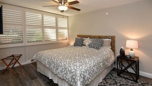 A bedroom with a bed, a ceiling fan, a window with blinds, a bedside table with a lamp, and a patterned rug. - Say Aloha to Island Life! Pool View Suite w/Kitchen Ease, WiFi–Kamaole Sands 6308 (KIHEI)