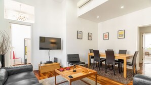Un salon moderne avec une table basse en bois, des canapés en cuir noir, un coin repas avec une table et des chaises en bois, un téléviseur à écran plat fixé au mur et des tableaux encadrés accrochés au mur.