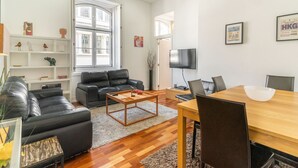 Un salon moderne avec un canapé en cuir noir, une table basse en bois et une table à manger avec des chaises. Un téléviseur est fixé au mur et une bibliothèque contient des objets décoratifs.