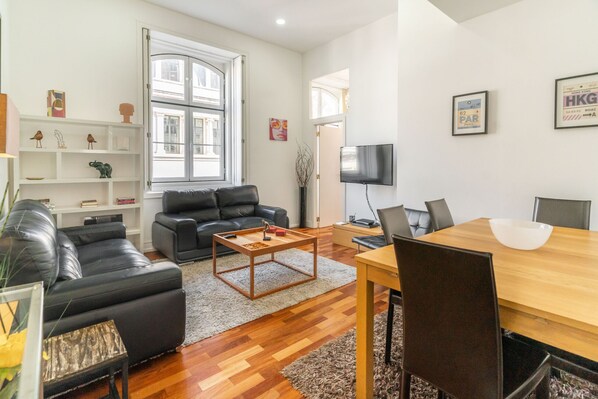 Un salon moderne avec un canapé en cuir noir, une table basse en bois et une table à manger avec des chaises. Un téléviseur est fixé au mur et une bibliothèque contient des objets décoratifs.