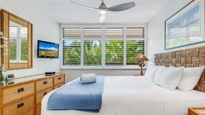 A bedroom with a bed, a ceiling fan, a TV, a window with blinds, and a framed picture on the wall.