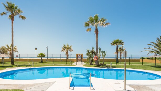 A swimming pool with a clear view of the ocean, surrounded by palm trees and a fenced area. - Beachfront apartment in Tarifa with pool (Tarifa)
