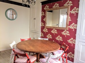 A dining area with a round wooden table surrounded by four chairs, a wall-mounted round mirror, and a red floral wallpaper.