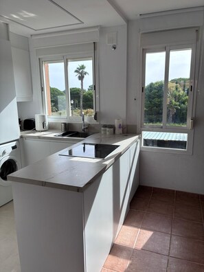 A modern kitchen with a central island, a built-in washing machine, and two windows with views of greenery.