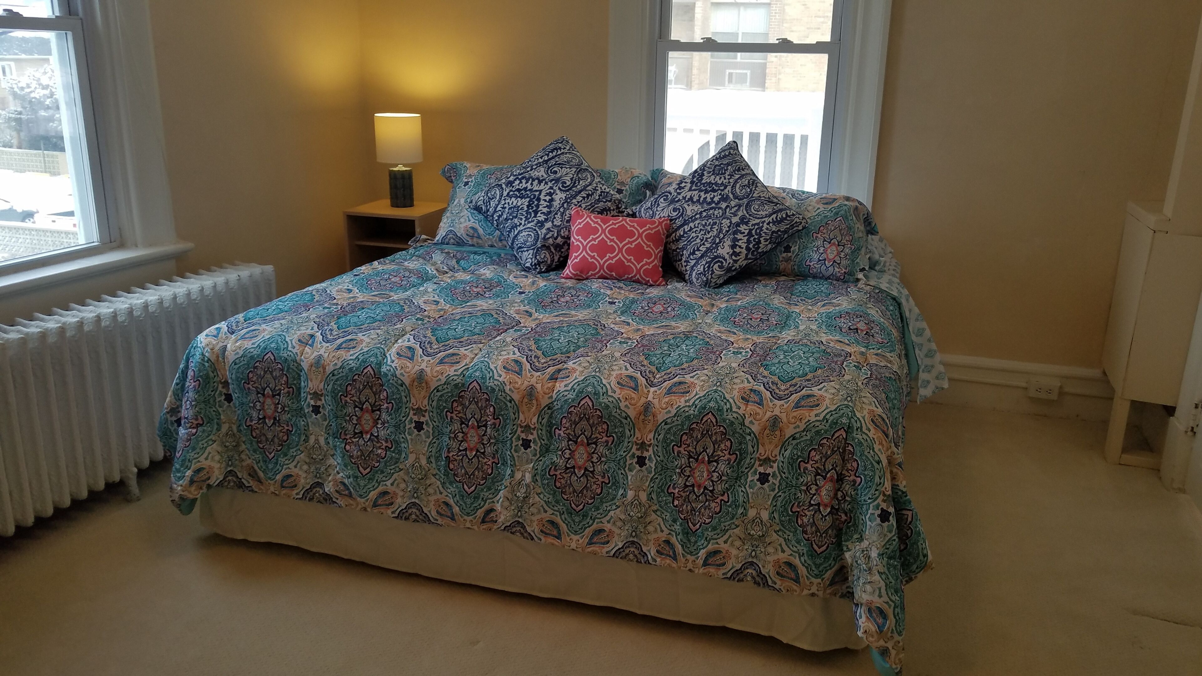 Un dormitorio con una colcha estampada, una mesita de noche con lámpara y una ventana con vistas al exterior.