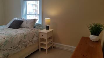 A bedroom with a bed, a bedside table with a lamp, a small plant, and a window with a view of buildings.
