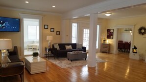 A spacious living room with hardwood floors, a dark-colored sofa, a white ottoman, and a television on the wall. - Beach Block 11 BDR Home (Ventnor City)