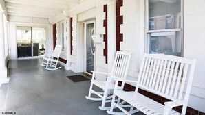 A covered outdoor seating area with white rocking chairs and benches, large windows, and a concrete floor. - Beach Block 11 BDR Home (Ventnor City)