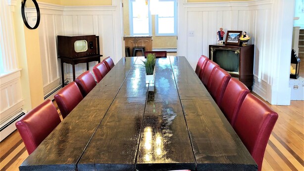 A dining room with a long wooden table, red upholstered chairs, a small potted plant, and a vintage television set. - Beach Block 11 BDR Home (Ventnor City)