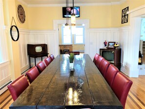 A dining room with a long wooden table, red upholstered chairs, a small potted plant, and a vintage television set. - Beach Block 11 BDR Home (Ventnor City)