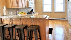 A kitchen with wooden cabinets, a granite countertop, a bowl of fruit, and three stools. - Beach Block 11 BDR Home (Ventnor City)