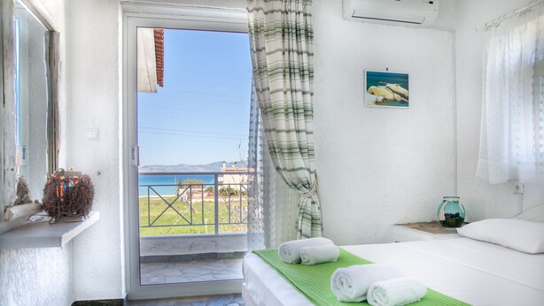 A hotel room with a bed, a green towel, a small table with a vase, and a view of the sea through a door. - Independent condo, with stunning view just a heartbeat away from the beach (Kokkonion)