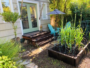 Eine Holzterrasse mit einer Bank, einem blauen Stuhl, einem Blumenbeet mit Pflanzen und einem Haus mit grüner Tür.