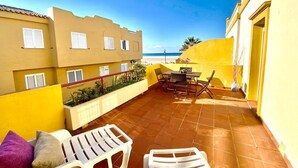 A terrace with a table and chairs, a white chair, a small table with a vase, and a view of the beach. - Beachfront apartment with large terrace (Tarifa)