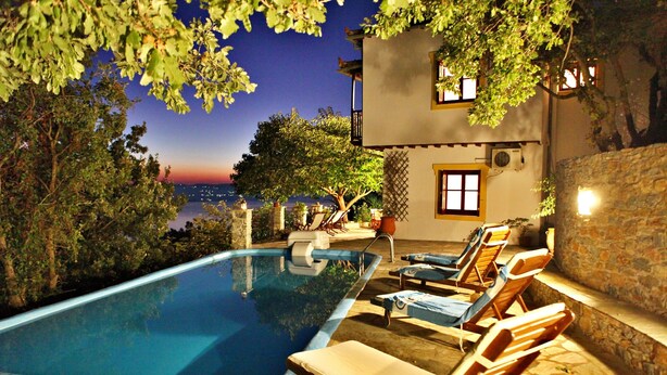 A swimming pool with lounge chairs, a stone wall, and a view of the sea at dusk.
