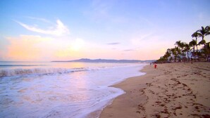 Una platja amb una persona caminant, palmeres i un capvespre.