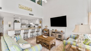 A spacious living room with a large sofa, a coffee table, and a kitchen area with white cabinets and a bar. - Unit 15, Pet Friendly 3 Bedroom Beach Condo (Panama City Beach)