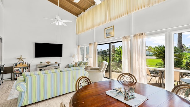 A spacious living room with a large sofa, a wooden dining table, and a television mounted on the wall. - Unit 15, Pet Friendly 3 Bedroom Beach Condo (Panama City Beach)