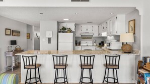 A modern kitchen with white cabinets, a granite countertop, and four bar stools. - Unit 15, Pet Friendly 3 Bedroom Beach Condo (Panama City Beach)