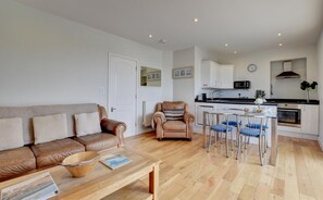 A compact living space with a sofa, a dining table with chairs, a kitchen area with a microwave, and a clock on the wall.