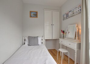 Une petite chambre blanche avec un lit simple, une commode, un miroir et un petit tabouret.