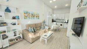 Uma sala de estar moderna com sofá bege, mesa de centro branca e área de cozinha com armários e eletrodomésticos brancos.