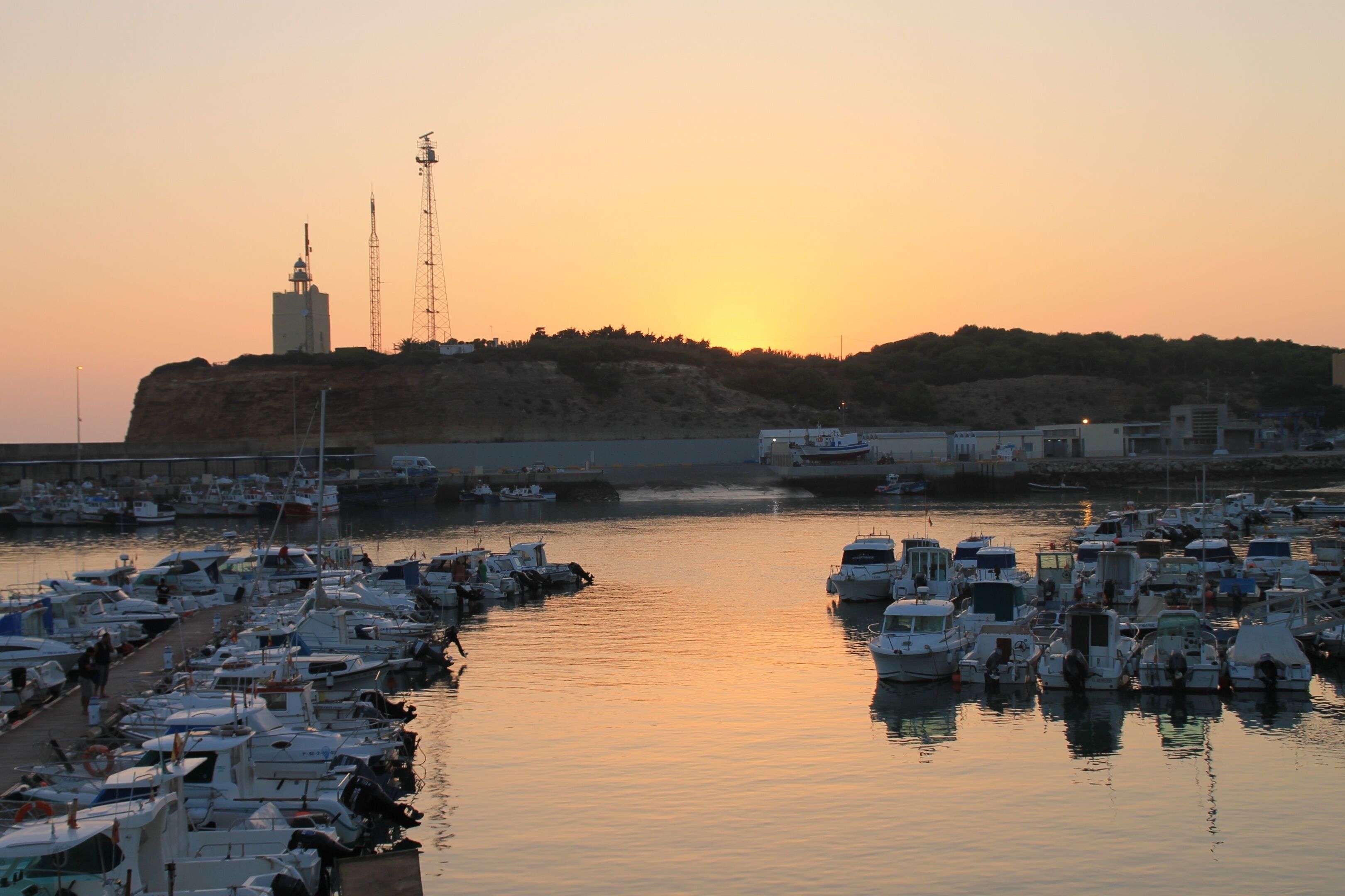 A marina with numerous boats docked, a prominent building with a tower, and a hilly landscape in the background.