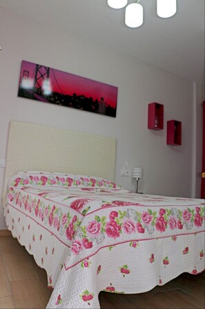 Ein Schlafzimmer mit geblümtem Überwurf, einem Stadtbild an der Wand und einem Nachttisch mit Lampe.
