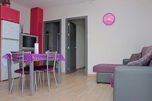 Ein kleiner, moderner Wohnraum mit Essbereich, einem grauen Sofa, einer pinken Uhr und einer Küchenzeile mit einem roten Schrank und einem weißen Kühlschrank.