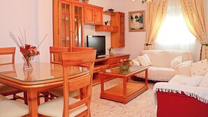 Un salon comprenant une table à manger avec un plateau en verre, des chaises en bois, un canapé, un téléviseur, des meubles en bois et un tableau accroché au mur.